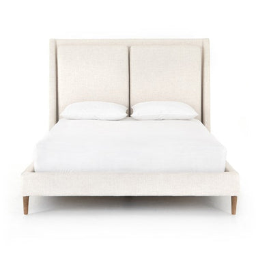 A Poppy Bed with a beige upholstered headboard and tapered wooden legs, with a mattress and two pillows on top.